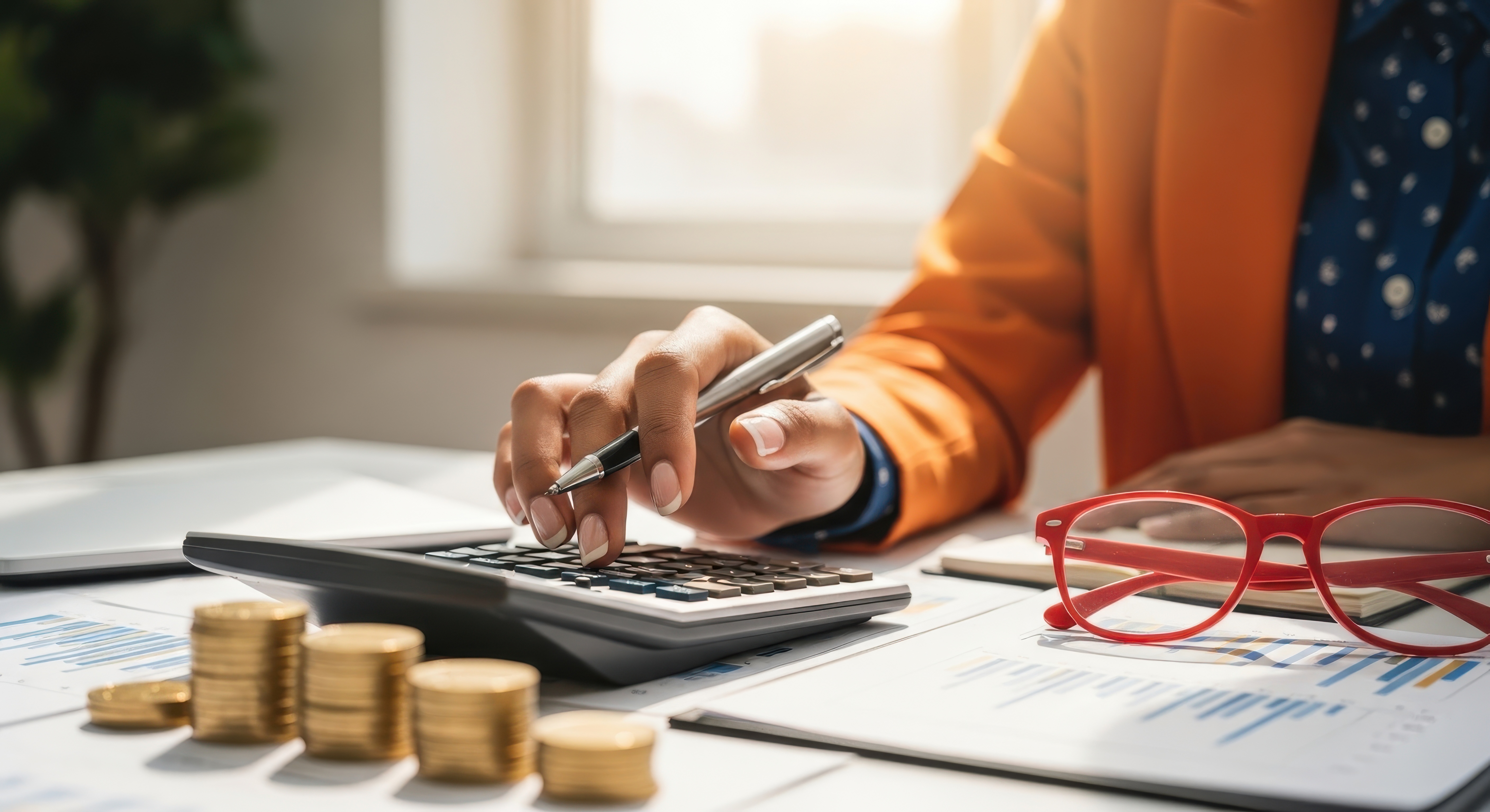 View of a person, dressed in an orange jacket, using a calculator with a pen in hand while seated at a desk with stacks of gold coins, financial documents showing charts, and a pair of red-framed reading glasses resting nearby, suggesting detailed financial work or bookkeeping.